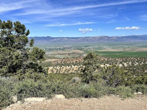 A panoramic view of a valley surrounded by mountains, with green fields and trees in the foreground under a blue sky.