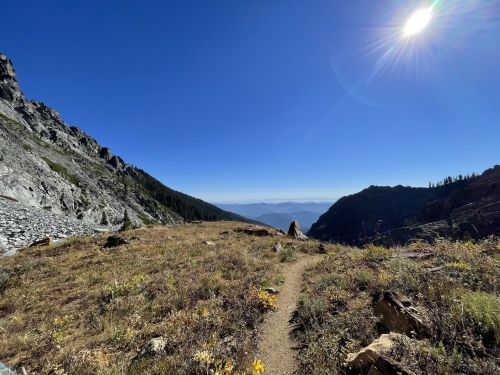 A scenic mountain trail with rocky terrain, surrounded by greenery and distant mountains under a clear blue sky.