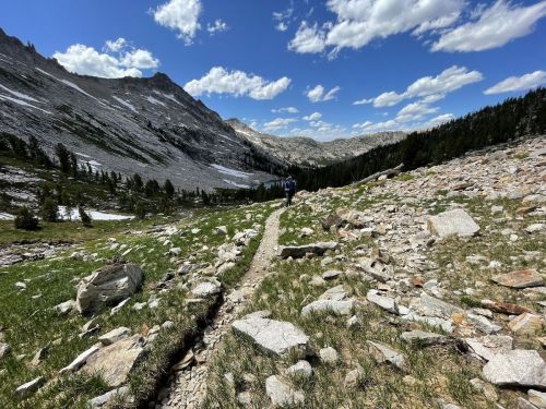 A rocky trail winds through a mountainous landscape under a blue sky with scattered clouds.