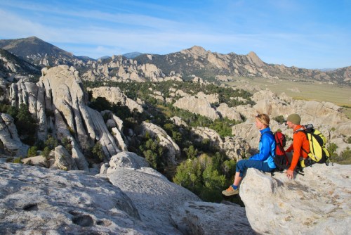 Two hikers sit on a rock ledge, overlooking a rugged landscape of boulders and mountains under a clear blue sky.