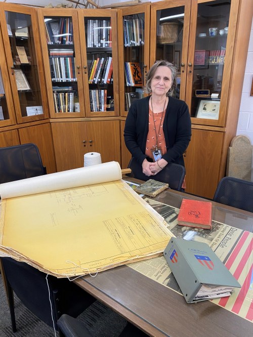 A woman stands beside a large blueprint on a table, with books and artifacts displayed in a library setting.