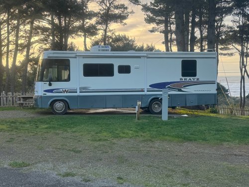 A parked RV named "Brave" near a grassy area, with trees and a coastal view in the background at sunset.