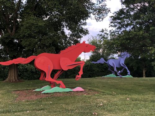Two colorful horse sculptures, one red and one purple, galloping on a grassy area with trees in the background.