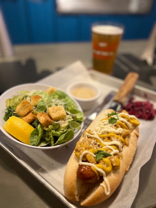 A hot dog topped with mustard and mayo, served with a side salad, croutons, and a drink on a tray.