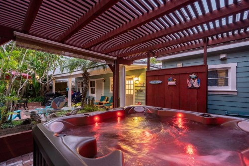 A hot tub with red lights under a wooden pergola, surrounded by tropical plants and a cozy backyard setting.