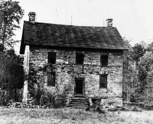 A historic stone house with a symmetrical facade, two stories, and a front staircase, surrounded by trees.