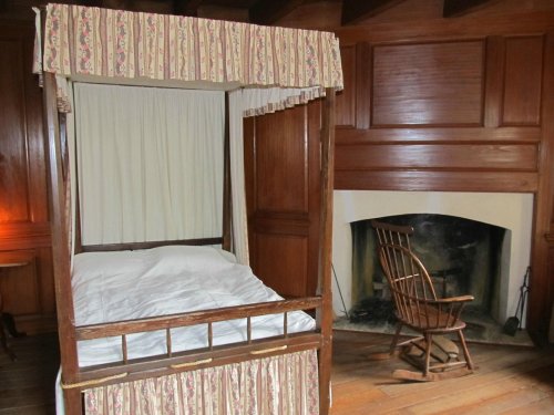 A cozy bedroom featuring a four-poster bed with curtains and a wooden rocking chair near a fireplace.