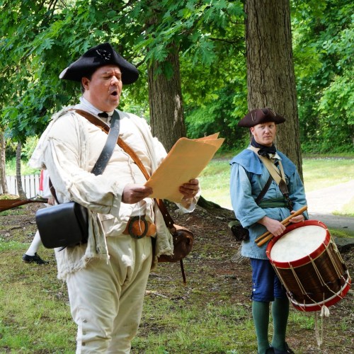 Two historical reenactors in colonial attire, one reading a document and the other playing a drum, surrounded by trees.