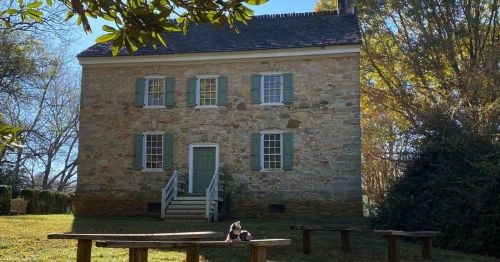 A historic stone house with green shutters, surrounded by trees and a grassy area, featuring a wooden bench in front.