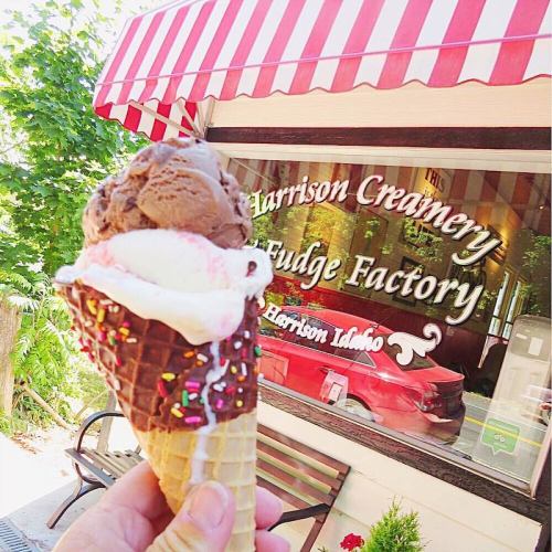 A hand holds a colorful ice cream cone in front of the Harrison Creamery and Fudge Factory window.