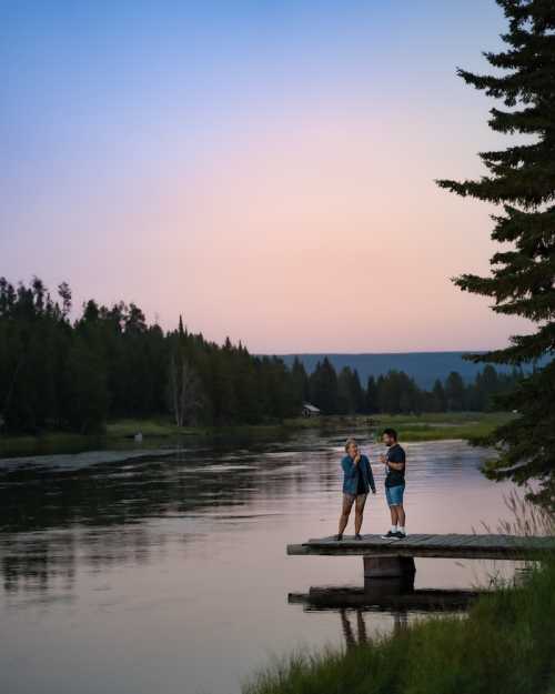 A couple stands on a wooden dock by a calm lake at sunset, surrounded by trees and mountains in the background.