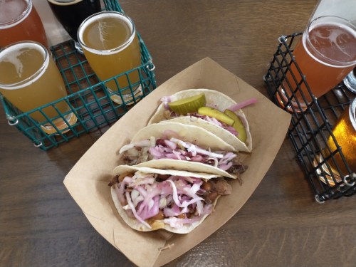 Three tacos with meat, pickles, and onions, served in a paper tray, alongside several glasses of beer.