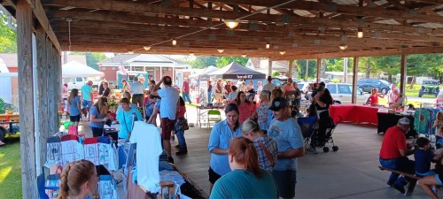 A bustling outdoor market scene with people shopping, chatting, and browsing various stalls under a wooden pavilion.