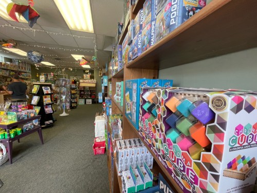 A colorful toy store aisle filled with games and puzzles, featuring shelves of vibrant products and a customer browsing.