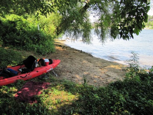 A red kayak rests on a sandy shore beside a calm river, surrounded by lush greenery and trees.