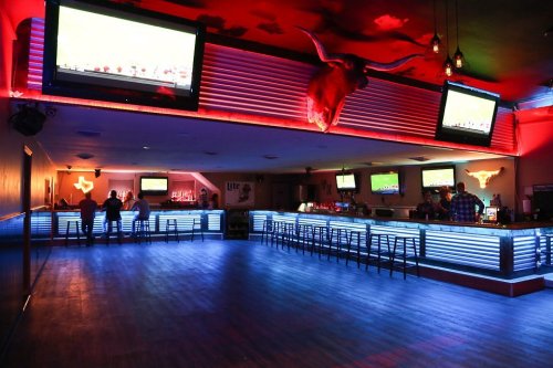 A dimly lit bar with blue LED lighting, multiple TVs showing sports, and a long counter with stools.