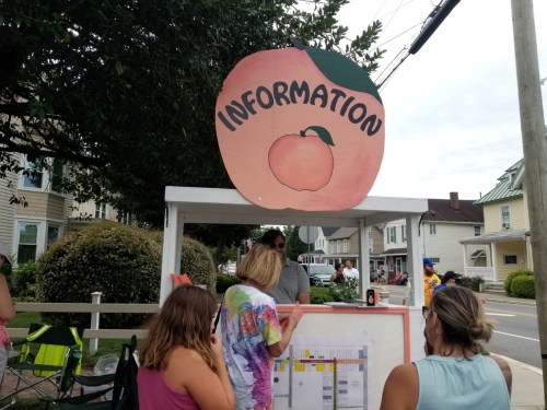 A booth with a peach sign labeled "INFORMATION" and people gathering around it on a sunny day.