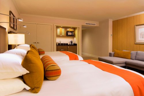 A cozy hotel room featuring two beds with orange accents, a seating area, and a kitchenette in the background.