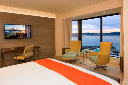 A modern hotel room featuring a bed, desk, TV, and two chairs with a view of the water and city skyline.