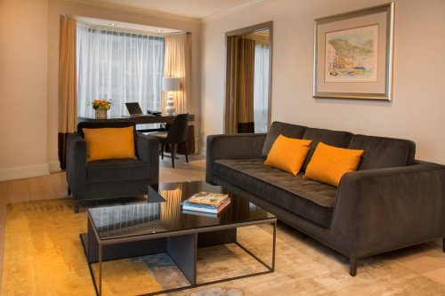 A modern hotel room featuring a gray sofa with orange pillows, a glass coffee table, and a workspace by a window.