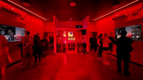 A dimly lit room with red lighting, featuring people interacting with screens on the walls.