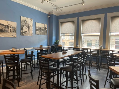 A cozy restaurant interior with blue walls, wooden tables, and large windows, featuring black chairs and framed photos.