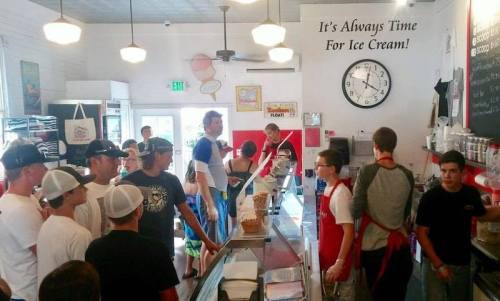 A busy ice cream shop with a line of customers and staff behind the counter, featuring a clock and a sign about ice cream.