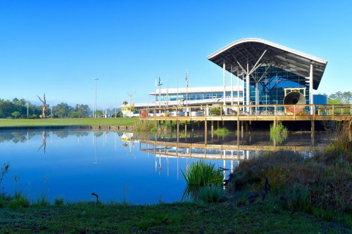 A modern building with a curved roof reflects in a calm lake, surrounded by greenery and clear blue skies.