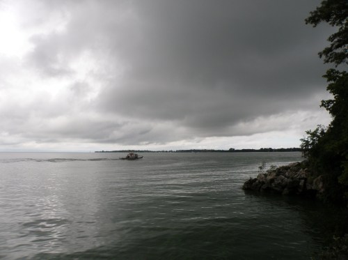 A boat glides across a calm lake under a cloudy sky, with trees and rocky shore visible in the foreground.