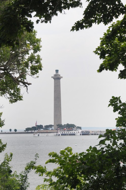 A tall monument stands on a shoreline, framed by green trees, with a calm body of water in the foreground.
