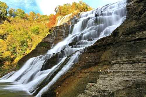 A cascading waterfall flows over rocky cliffs, surrounded by lush greenery and autumn-colored trees.