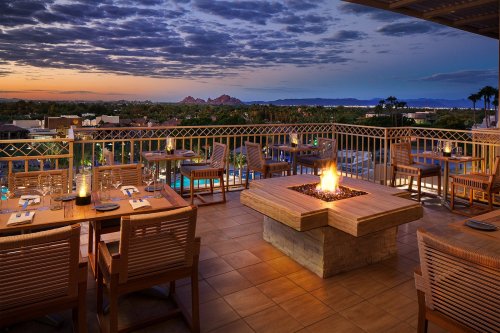 A cozy restaurant patio at sunset, featuring a fire pit, wooden tables, and scenic views of mountains and palm trees.