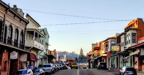 A quiet street lined with historic buildings and parked cars, bathed in warm morning light.
