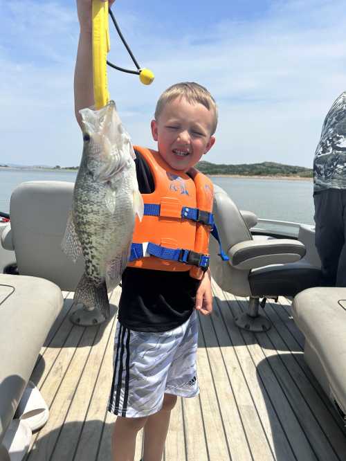 A smiling boy in a life jacket holds a large fish on a boat, with a lake and hills in the background.