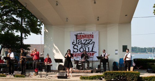 A band performs on stage at the Salem Jazz and Soul Festival, with a banner and waterfront in the background.