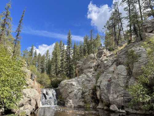 A serene waterfall cascades over rocks, surrounded by lush green trees under a bright blue sky with fluffy clouds.