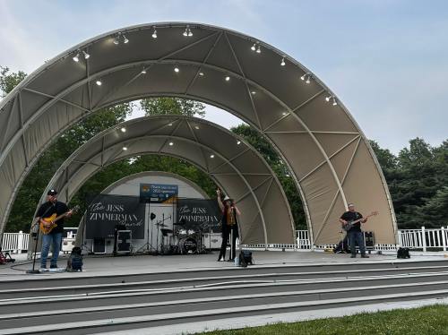 A band performs on stage under a large, curved canopy at an outdoor venue, surrounded by trees.