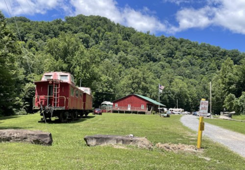 A red train caboose beside a red building, surrounded by green trees and a blue sky with clouds.