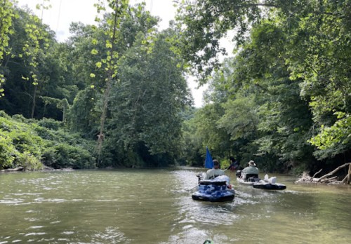 Two people on inflatable boats paddle down a serene river surrounded by lush green trees.