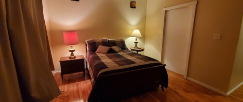 Cozy bedroom with a bed, two lamps, and warm lighting, featuring wooden floors and a closed door.