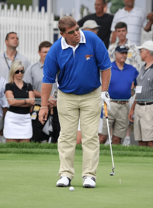 A golfer in a blue shirt and beige pants prepares to putt, surrounded by spectators on a sunny day.