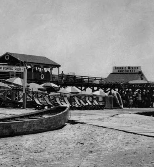 A black-and-white photo of a fishing pier with boats and beach chairs, bustling with people in the background.