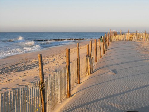 A sandy beach with wooden posts and fencing, gentle waves lapping at the shore under a clear sky.