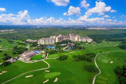 Aerial view of a luxury resort surrounded by lush greenery and golf courses under a blue sky with fluffy clouds.