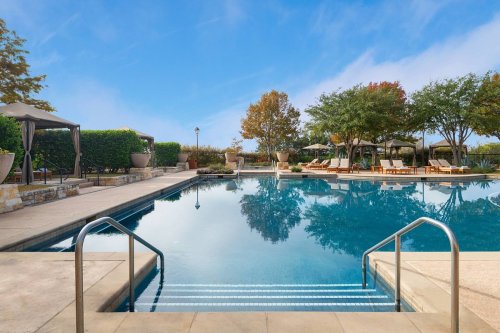 A serene pool area with lounge chairs, shaded cabanas, and trees reflecting in the water under a clear blue sky.