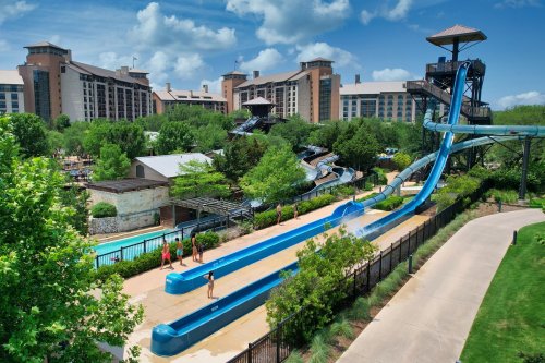 A water park with blue slides and a lazy river, surrounded by greenery and large resort buildings under a blue sky.