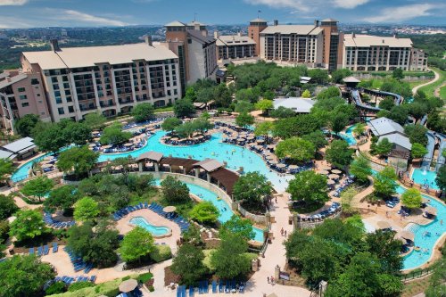 Aerial view of a resort with pools, water slides, and lush greenery, surrounded by large buildings under a blue sky.