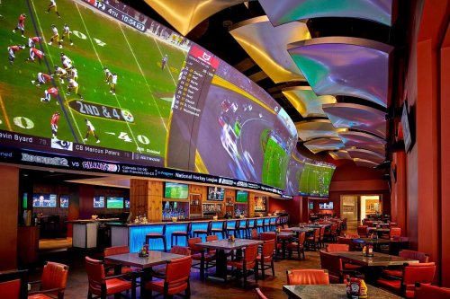 A spacious sports bar with large screens showing games, colorful lighting, and empty tables and chairs.