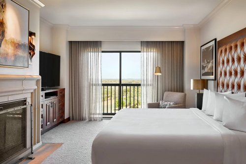 A modern hotel room featuring a king bed, armchair, TV, and large window with a view of greenery outside.