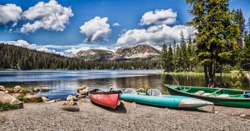 A serene lake scene with canoes on the shore, surrounded by trees and mountains under a blue sky with fluffy clouds.
